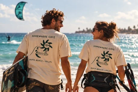 Two kitesurfers in matching Somewhere Café t-shirts walking along the shore with their boards