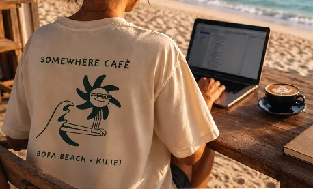 Someone working on a laptop at a beach table in a Somewhere Café t-shirt, with a latte beside them